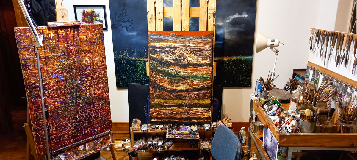 James de Villiers artist studio. Two abstract paintings in shades of brown, with many paint brushes and tubes of oil paint.
