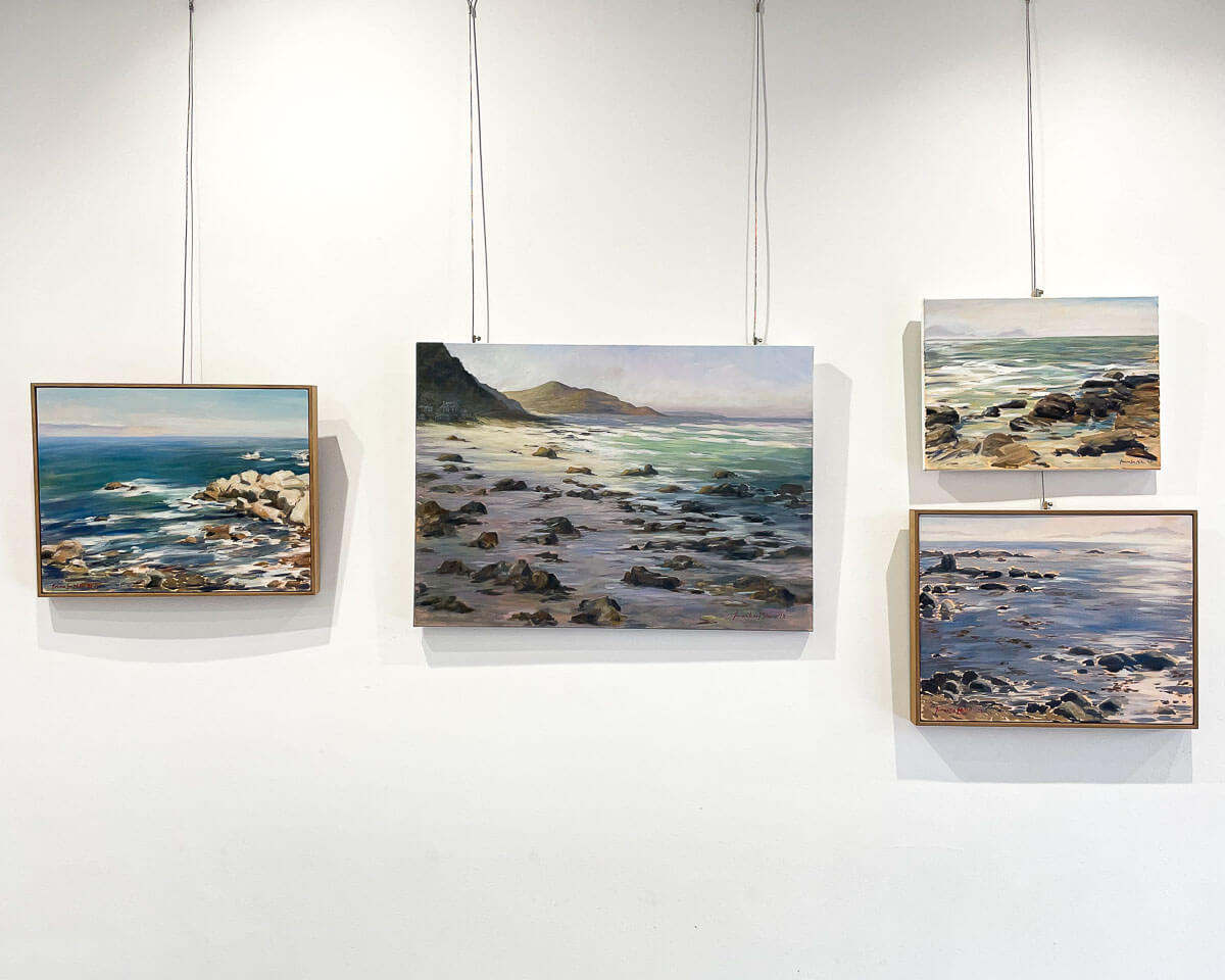Four paintings by Joanna Miller hanging on a white wall. The paintings all depict the ocean, with rocks and waves, painted en plein air.