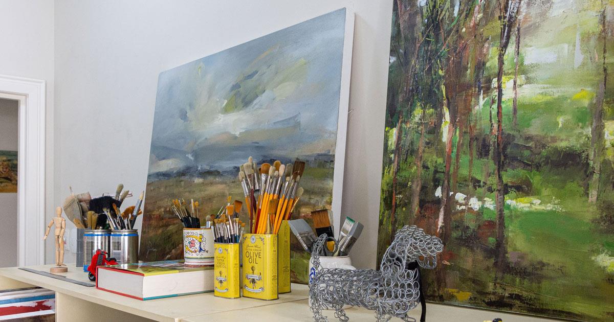 Janet Dirksen's art studio. There are 2 landscape paintings and her collection of paintbrushes and art materials arranged on the shelf.